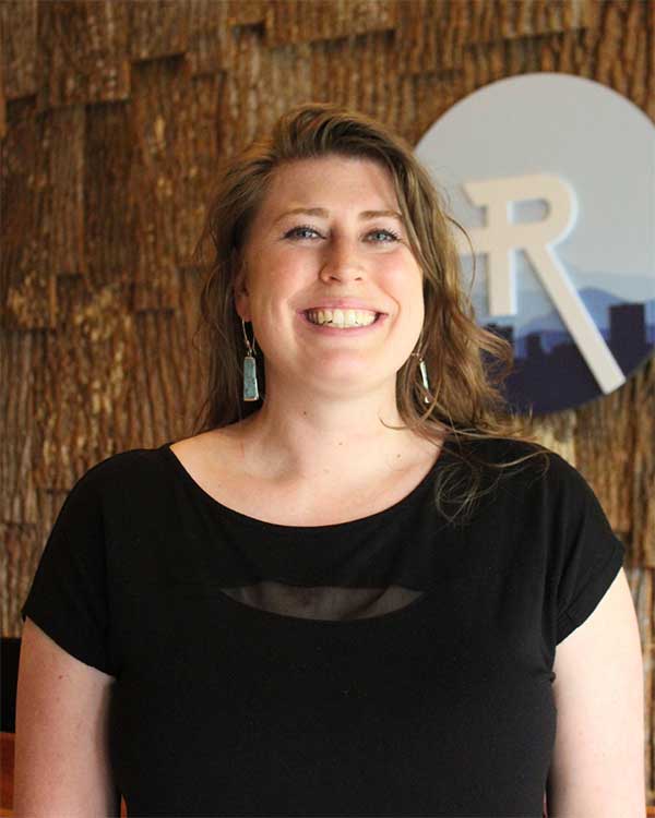 Dr. Sarah Ruszel, chiropractor at Radius Chiropractic, smiling warmly in a black top, with a natural wood background and clinic logo, reflecting her cheerful and empathetic approach to holistic health.
