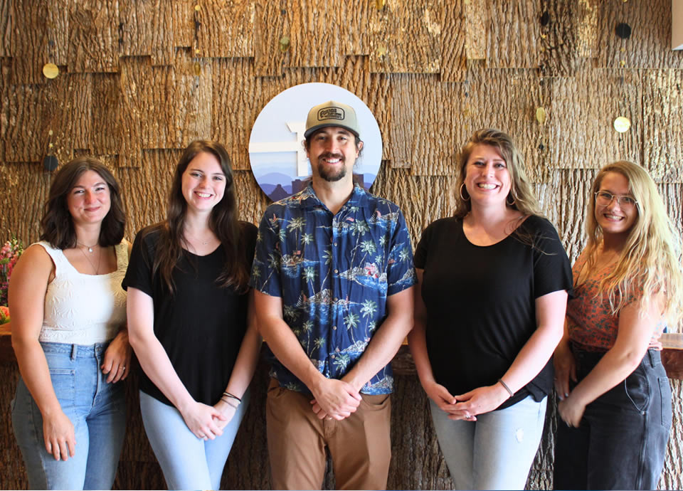 Team members of Radius Chiropractic posing in front of a textured wall with a logo, showcasing a friendly and welcoming atmosphere for the Asheville chiropractic practice.